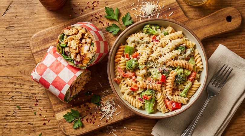 Chicken wrap in red checkered paper next to a bowl of pasta salad with broccoli and peppers.