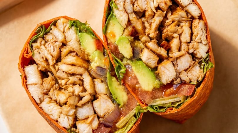 Two halves of a chicken wrap with avocado, lettuce, and tomatoes on a brown surface.